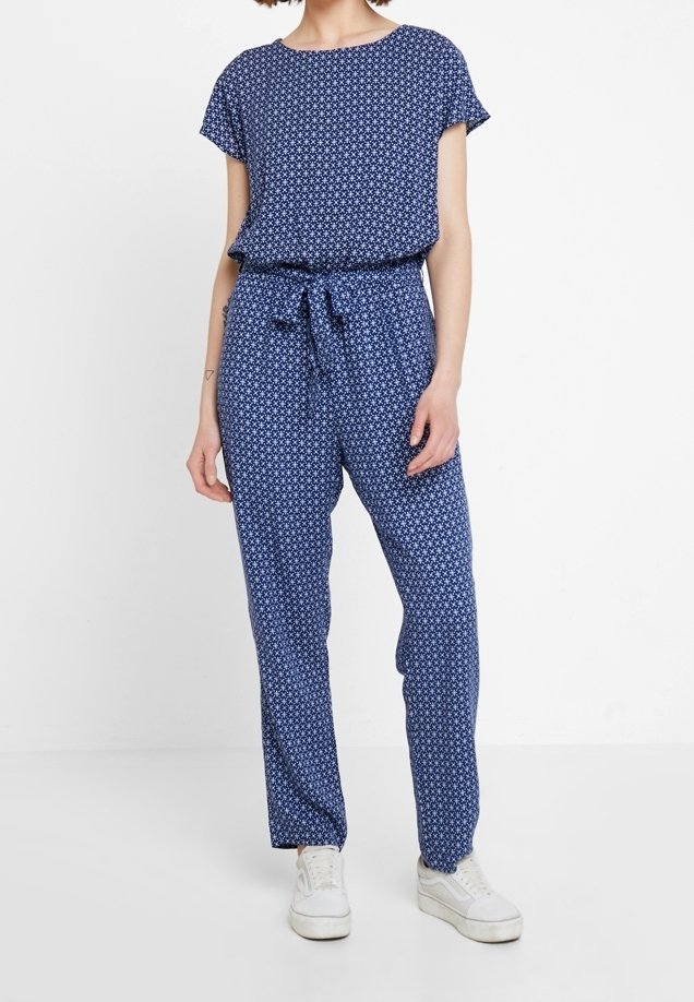 Femme portant une combinaison bleue à manches courtes avec un motif d'étoiles blanches et une taille nouée, associée à des baskets blanches, debout devant un fond blanc.
