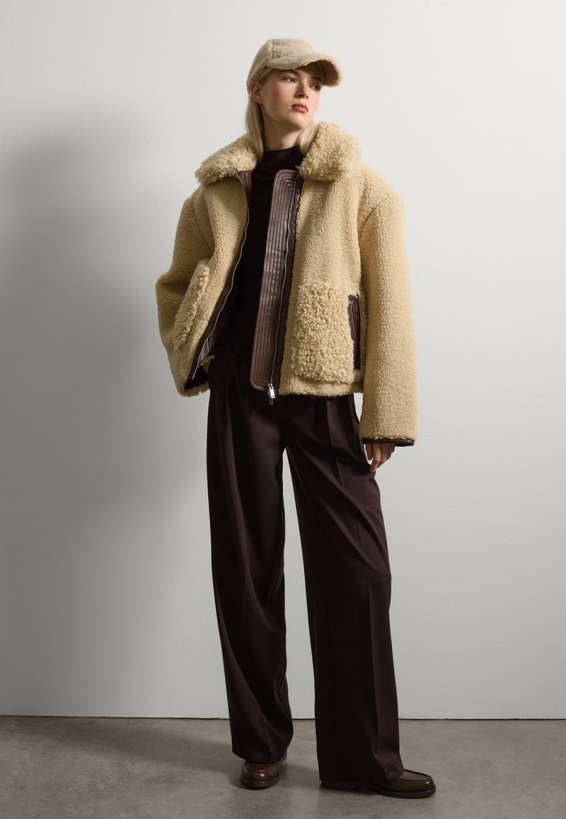 Giacca in pelliccia sintetica beige con zip frontale, ampio colletto e dettagli marroni, abbinata a pantaloni larghi marrone scuro e un cappello beige.