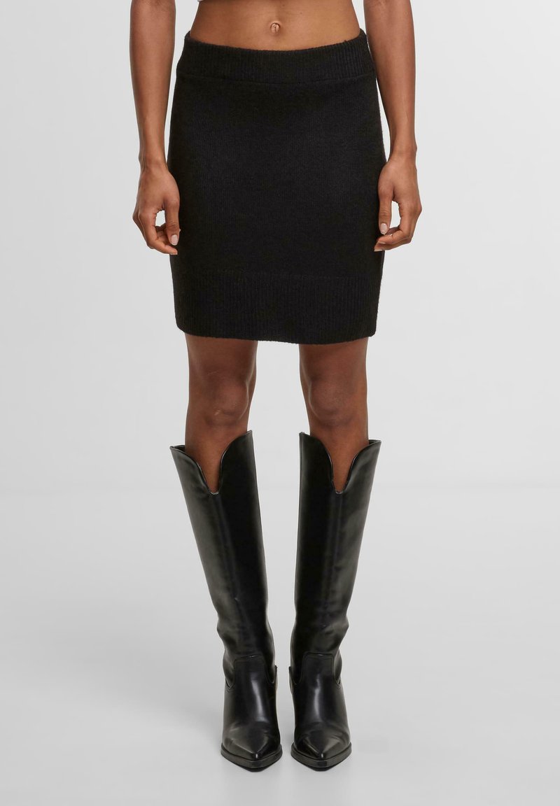 Černá pletená sukně s přiléhavým střihem, žebrovaným páskem a lemem. Vytvořeno ve spojení s vysokými černými koženými botami na špičatou špičku, které mají široké otevření.