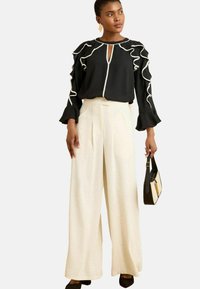Blusa nera con dettagli in raso bianco e scollatura a chiave, abbinata a pantaloni ampi crema e una piccola borsa nera.