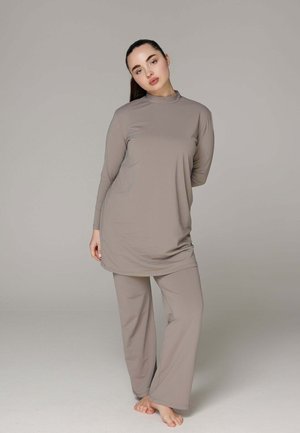 Femme aux cheveux foncés posant pieds nus, portant une tunique à manches longues ample taupe et un pantalon large assorti devant un fond gris uni.