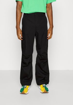 Personne portant un pantalon cargo noir, une chemise vert vif et des baskets jaune-vert-blanc, debout devant un fond clair uni.