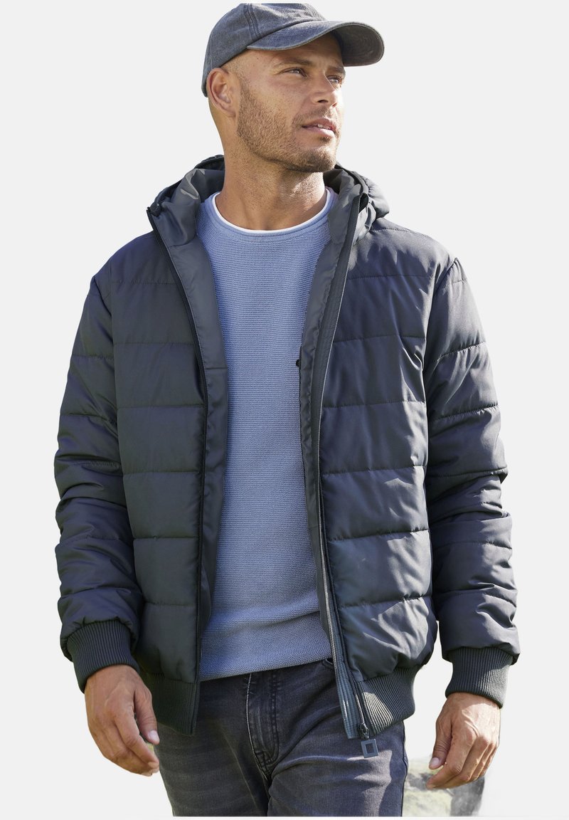Giubbotto imbottito grigio scuro, con cappuccio, polsini a costine e chiusura con zip. Indossato sopra un maglione azzurro chiaro e abbinato a jeans.