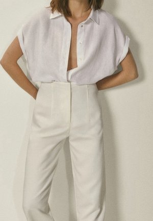 Femme portant une chemise blanche ample à manches courtes et boutons, rentrée dans un pantalon blanc taille haute, se tenant les mains sur les hanches devant un fond uni.