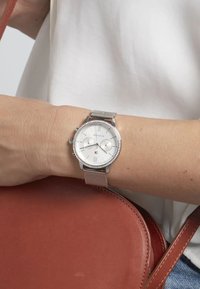 Orologio con cinturino in rete argentata, quadrante rotondo bianco, dettagli argentati e pietre decorative sulla lunetta. Una mano poggia su una borsa in pelle rossa.