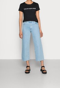 Černé tričko s krátkým rukávem a bílým logem "CALVIN KLEIN JEANS", spárováno se světlými modrými džínami s vysokým pasem a širokými nohavicemi a černými sandály.