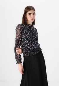 Blusa nera floreale in tessuto trasparente, con orlo arricciato, maniche lunghe con polsini elasticizzati e dettagli a oblò sul neckline.