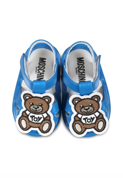 Sandales pour bébé en synthétique bleu avec un design à découpes, présentées avec une patch en forme d'ours en peluche étiqueté "Jouet" sur chaque chaussure. Fermeture à patte Velcro.