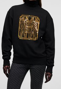 Felpa nera con un design geometrico dorato e il logo "Karl Lagerfeld"; presenta polsini e colletto a coste, abbinata a jeans neri con fantasia.