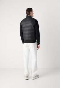 Μαύρο πλισέ γιλέκο με ψηλό γιακά, φορεμένο πάνω από μαύρη μπλούζα με μακριά μανίκια, συνδυασμένο με λευκά παντελόνια. Ελαφρύ υλικό και στρωτή σχεδίαση.