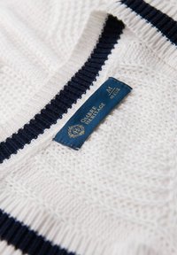 Maglione bianco lavorato a maglia con collo a coste blu navy e dettagli a righe. Presenta un'etichetta con la scritta "Ombre Heritage", taglia M, con una texture decorativa.