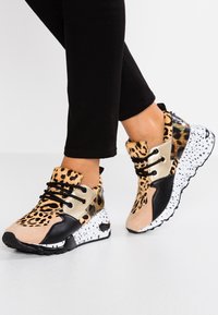 Personne portant un imprimé léopard, des baskets noires et beige avec des lacets noirs et des semelles blanches tachetées, se tenant sur un sol blanc avec un pantalon noir.