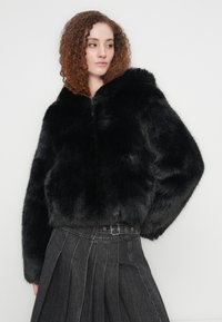 Γυναίκα με σγουρά κόκκινα μαλλιά φοράει μαύρο σακάκι από γούνα και ψηλόμεση σκουρόχρωμη γκρι φούστα με πιέτες, ποζάροντας πλάγια μπροστά σε απλό φόντο.