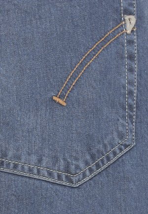 Gros plan sur un tissu en denim bleu avec une poche arrière cousue présentant des lignes de couture doubles orange et blanche et un petit rivet métallique.