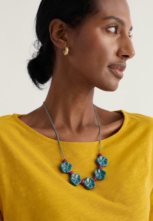 Turkist blomsterformet halskæde med røde perler på en blå snor, båret over en gul skjorte med en bred halsudskæring. Glat tekstur og levende farver.