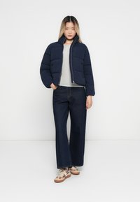 Ung kvinne med en marineblå pufferjakke, lys grå skjorte, vide mørke jeans og beige mønstrede joggesko som står mot en hvit bakgrunn.