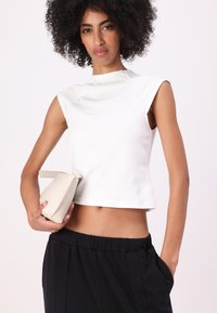 Baltas bezpiedurkņu crop top ar augstu kakla izgriezumu, savienots ar melniem vaļīgiem biksēm. Modelis tur nelielu, noapaļotu bēšā krāsā roku somu.