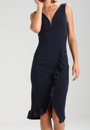 Robe midi sans manches bleu marine avec un décolleté profond en V et un ourlet asymétrique à volants portée par une femme avec la main près de son visage.