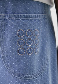 Denimplagg med et lyseblått stoff med prikkete mønster. Inkluderer en baklomme dekorert med brune broderte sirkler.
