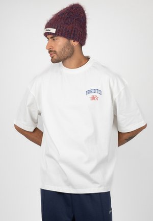 Homme portant un t-shirt blanc oversize avec le texte "PROHIBITED" et un bonnet en tricot bordeaux, regardant sur le côté avec les mains dans le dos.