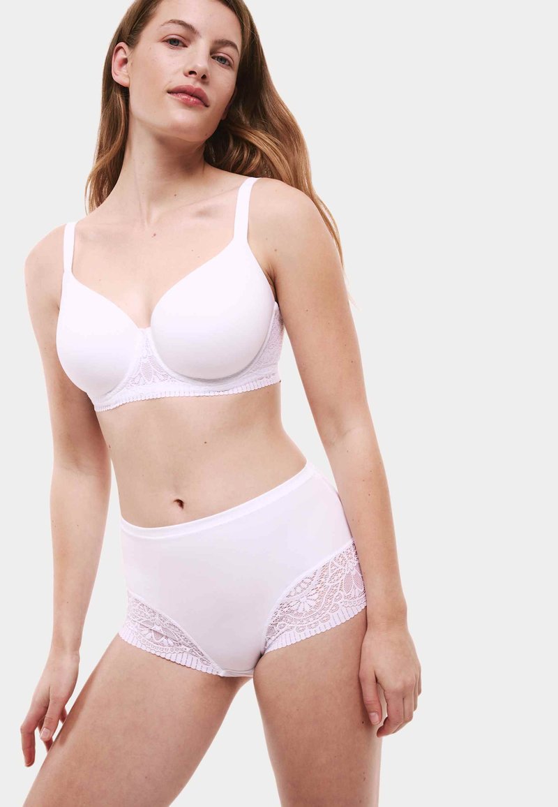 Donna che indossa un reggiseno bianco e mutandine a vita alta con dettagli in pizzo, in piedi contro uno sfondo chiaro e semplice, rivolta verso l'obiettivo.