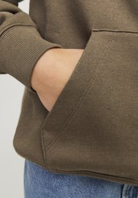 Sudadera marrón con puños acanalados, mostrando una mano en el bolsillo delantero. La tela tiene una textura suave y se ajusta alrededor de la cintura.