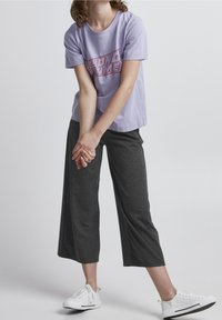 Světle fialové bavlněné tričko s grafickým nápisem "BEYOND TIME" v červené barvě, doplněné o černé zkrácené kalhoty a bílé tenisky s černými akcenty.