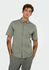 Chemise à manches courtes, boutonnée, de couleur olive, avec un col évasé, une texture subtile et des boutons foncés. Portée avec un pantalon gris à motifs.