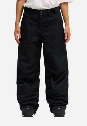 Person trägt lockere schwarze Hose, weißes Hemd und schwarze Schuhe, steht vor einem schlichten weißen Hintergrund.