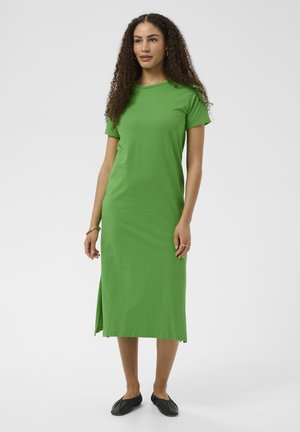 Moteris su garbanotais plaukais, vilkinti žalią midi su trumpomis rankovėmis su šoniniu plyšiu ir juodais lygiais batais, stovi su neutraliu veido išraiška.