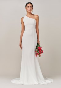 Vestido blanco de un solo hombro con una silueta ajustada, falda hasta el suelo y cola. La modelo sostiene un ramo de flores rosas. Textura suave.