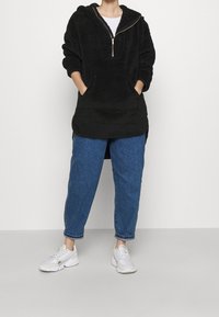 Person iført sort fuzzy hættetrøje med forlommer, blå opbrettede jeans og hvide sneakers, stående foran en neutral baggrund.