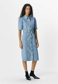 Vestido midi de mezclilla en azul claro, con botones en el frente, mangas cortas abullonadas, cintura ceñida con cinturón, bolsillos laterales, combinado con tacones negros y calcetas.