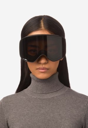 Zwarte skibril met een dikke rand en een oversized lens, met een matte afwerking. Draagt men boven een grijze coltrui.