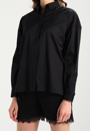 Camicia nera con bottoni realizzata in tessuto morbido, caratterizzata da un colletto classico, maniche lunghe e una vestibilità rilassata, abbinata a pantaloni corti in pizzo neri.