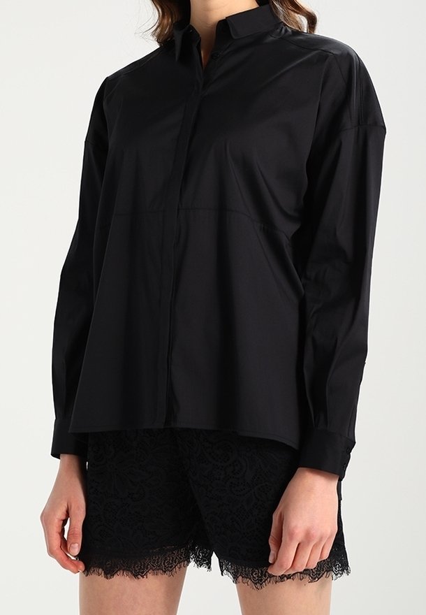 Camicia nera con bottoni realizzata in tessuto morbido, caratterizzata da un colletto classico, maniche lunghe e una vestibilità rilassata, abbinata a pantaloni corti in pizzo neri.