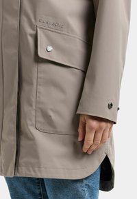 Beige vedenpitävä takki, jossa sivutasku ja nepparikiinnitys. Mukana sileä rakenne ja hillitty brändäys rintanevassa.