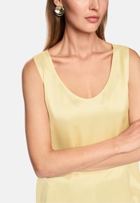 Yellow sleeveless top in smooth fabric with a deep neckline, paired with round metallic earrings. Skin tone visible on the arm.