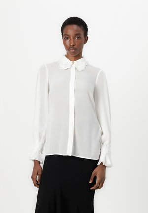 White blouse made of lightweight fabric, featuring a high collar with floral embellishments and button closure, long sleeves with ruffled cuffs.