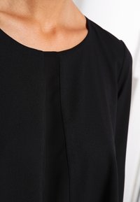 Blusa nera con scollatura arrotondata, caratterizzata da una cucitura frontale e una texture liscia e morbida. Il tessuto ha una leggera lucentezza.