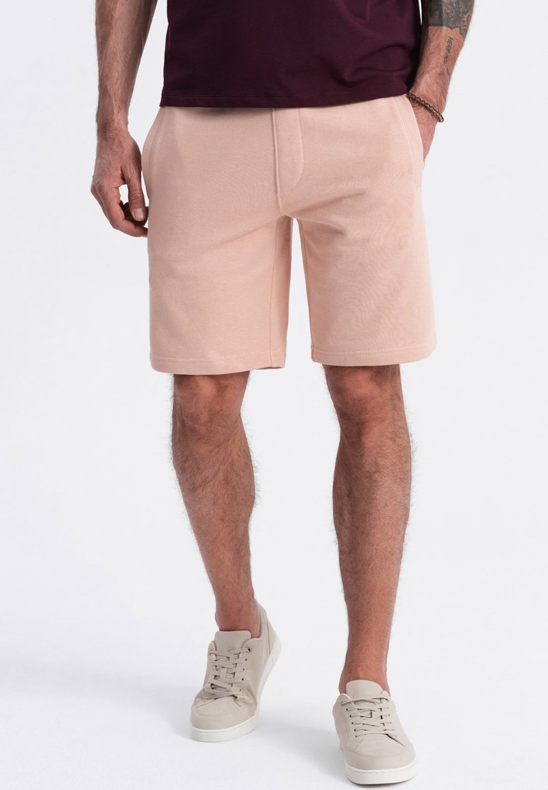 Shorts en coton pêche avec une coupe décontractée, dotés de poches latérales et d'une texture lisse, associés à des baskets beiges pour un look coordonné.