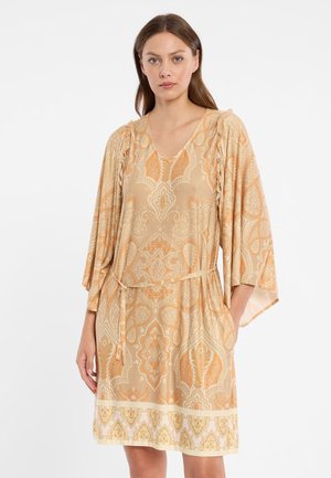 Femme portant une robe beige et orange à motifs, longueur genou, avec des manches trois-quarts et une fine ceinture à la taille, debout devant un fond blanc.