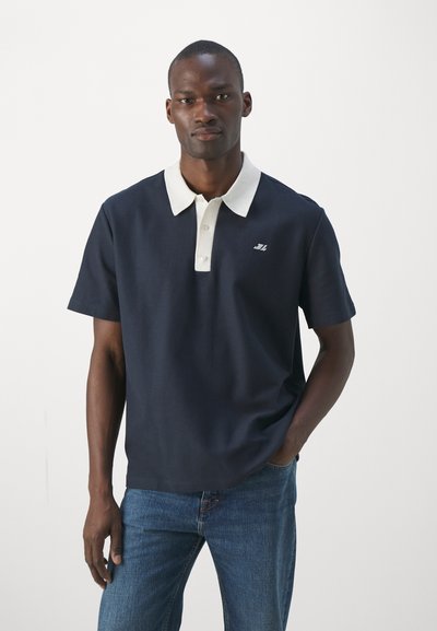 Marineblå polotrøye med hvit krage, korte ermer og logo på venstre side; kombinert med blå denimjeans.