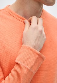 Μπλουζάκι μακρυμάνικο πορτοκαλί με υφή βάφλας, στρογγυλή λαιμόκοψη και ριμπ μανσέτες. Ένα χέρι κρατά απαλά το γιακά.