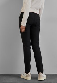 Černé slim-fit kalhoty se zadními kapsami, spárované s bílými teniskami se zlatými akcenty. Měkká, elastická látka s hladkým povrchem.