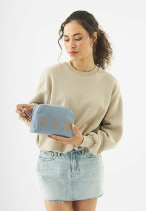 Frau in beigem Pullover und Jeansrock hält eine hellblaue Tasche mit drei Snoopy-Charakteren.