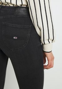 Jeans de denim negro con un bolsillo trasero curvado, que presenta un pequeño parche con el logo. La tela tiene una textura suave y un diseño ajustado.