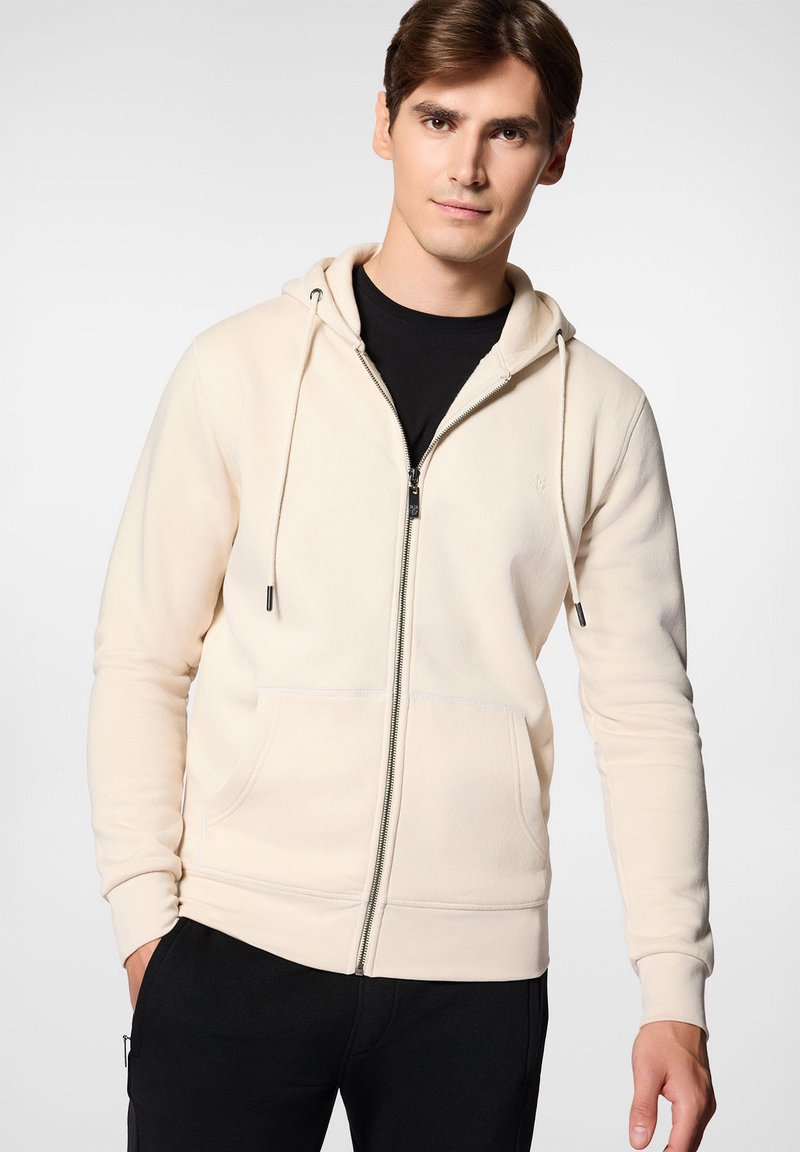 Kremowa bluza z kapturem na zamek, wykonana z miękkiego materiału, z kapturem na sznurki, kieszeniami z przodu i ściągaczami na rękawach. Nosi się ją na czarnej koszulce.