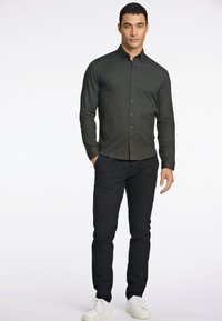 Camisa de botonadura en verde oscuro con un diseño ajustado, mangas largas y una textura suave, combinada con pantalones negros y zapatillas blancas.