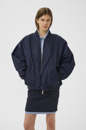 Giacca bomber blu navy realizzata in materiale leggero, con zip frontale, polsini a coste e spalle ampie e strutturate. Abbinata a una gonna a righe.